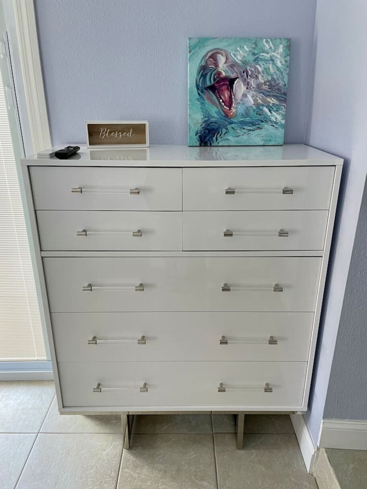 A white chest of drawers with multiple drawers, a remote control, and a framed artwork on top.