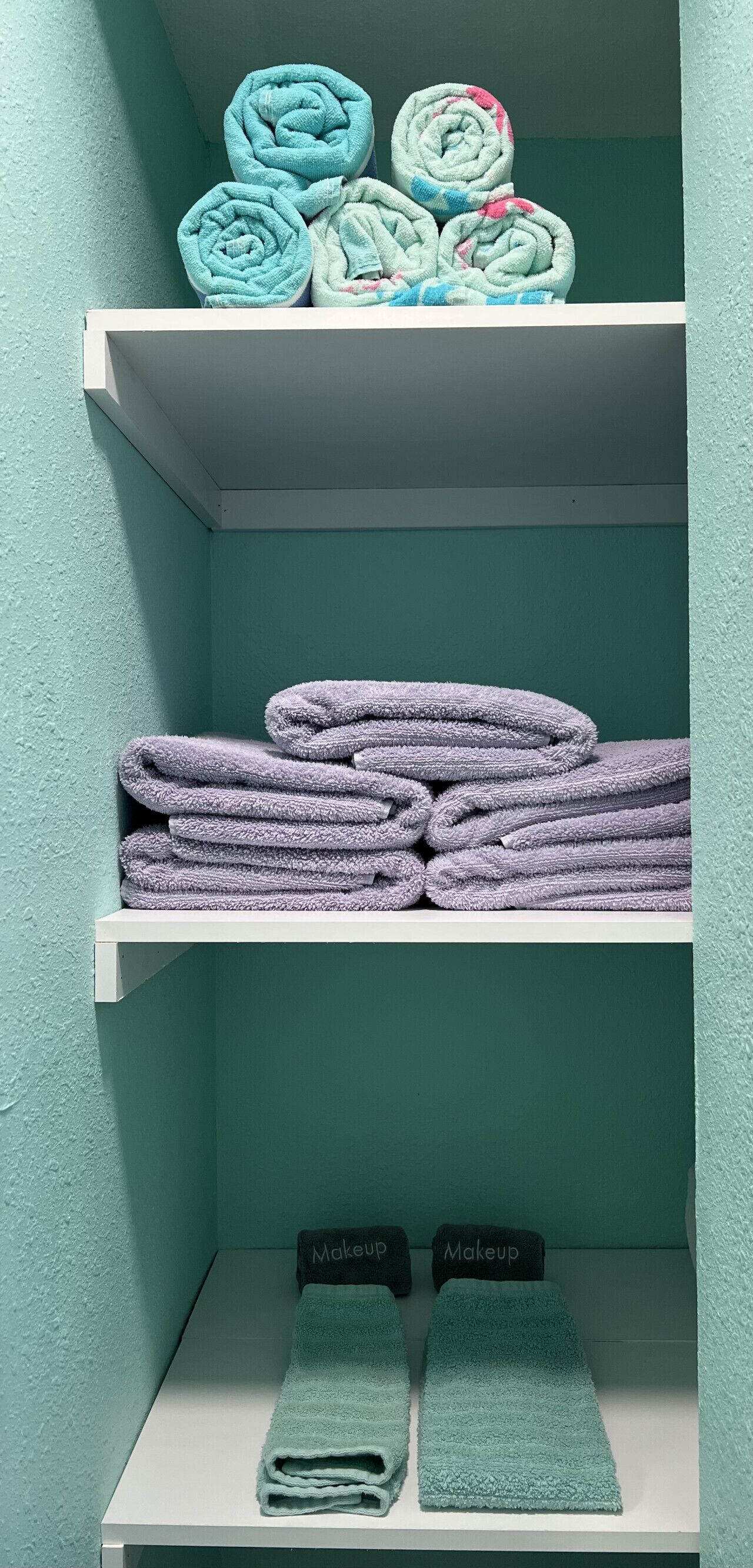 A bathroom with teal walls, a white shelf holding rolled towels, and two towels labeled "Makeup" on the floor.