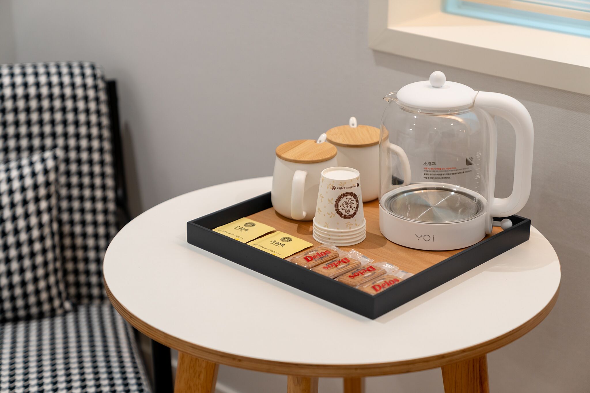 A small round table with a white coffee maker, two white mugs, a glass carafe, and a tray with tea bags and packets.