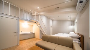 A modern hotel room with a wooden floor, a beige sofa, a wooden table, a bed with white linens, a staircase, and a bathroom with a sink and mirror. - Mono Hotel (Suwon)