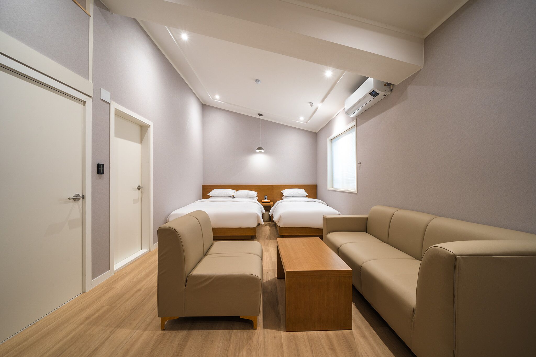 A modern hotel room with two beds, a sofa, a wooden coffee table, and an air conditioning unit on the wall.