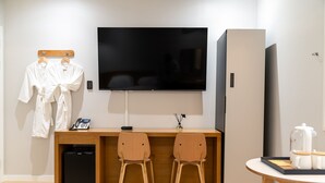 A hotel room with a wooden desk, two chairs, a TV, a minibar, and a small table with a kettle and cups. - Mono Hotel (Suwon)