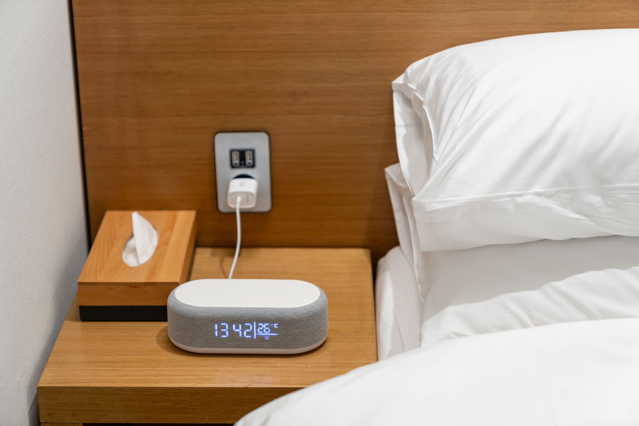 A bedside table with a digital clock displaying the time and temperature, a power outlet, and a tissue box.