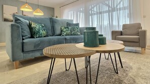 Una sala de estar moderna con un sofá azul, una mesa de centro redonda y un sillón gris. Hay dos lámparas colgantes sobre el sofá y un ventanal con cortinas translúcidas.