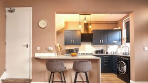 Une cuisine compacte avec des meubles de couleur sombre, un îlot central doté de tabourets, un lave-linge et une horloge au mur.