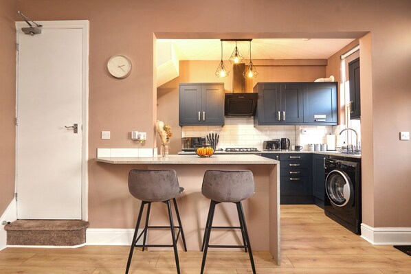 A compact kitchen with dark cabinets, a central island with stools, a washing machine, and a clock on the wall. - Homebird Property - Haddon House (Burley)