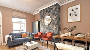 Un salon avec un canapé bleu, des fauteuils orange, une table basse blanche et un coin repas avec une table en bois mise pour quatre personnes.