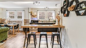 Eine moderne Küche mit einer zentralen Kochinsel, schwarzen Barhocker, einem Essbereich mit einem grünen Sofa und großen Fenstern mit Vorhängen.
