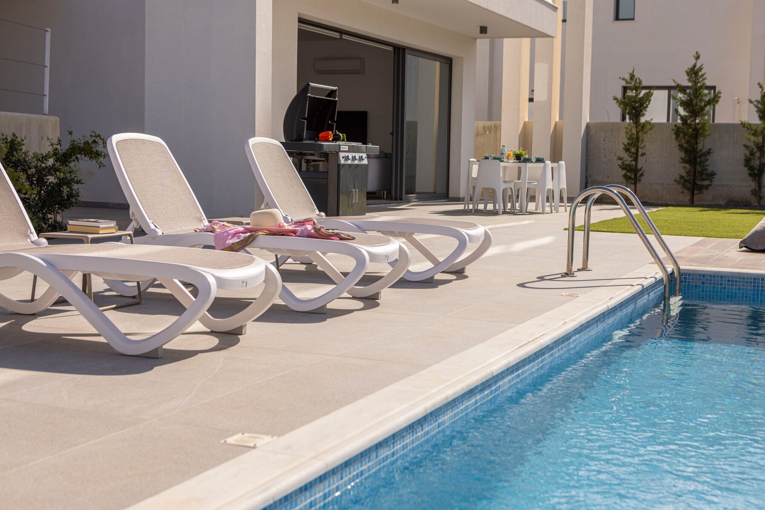 A poolside area with lounge chairs, a barbecue grill, and a small table with chairs.