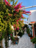 Un estrecho callejón con un edificio blanco en un lado, una mesa y sillas dispuestas afuera, y una maceta colgante con flores.