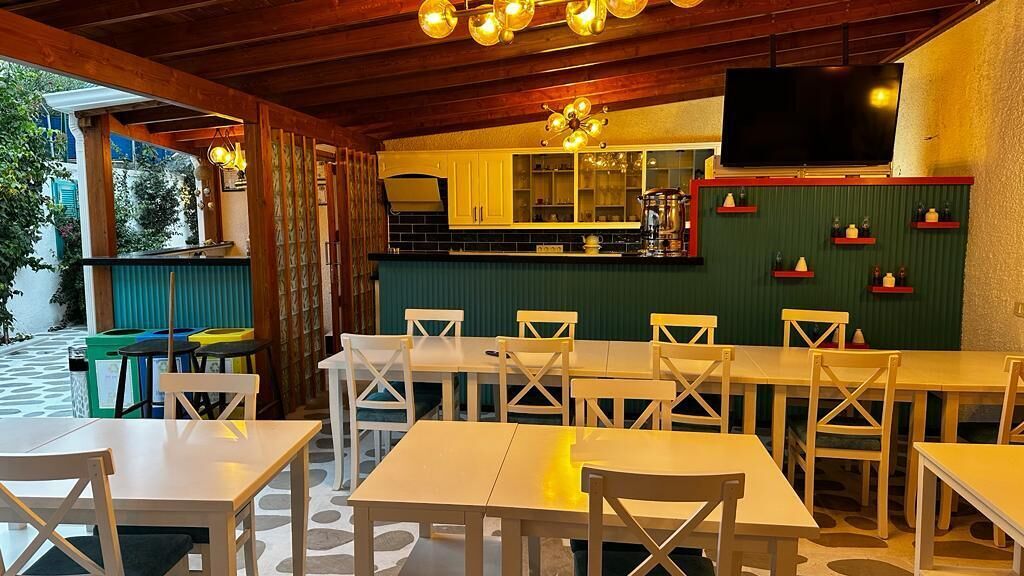 Un área de comedor al aire libre con mesas y sillas de madera, una pared verde con estantes y un televisor montado sobre la barra.