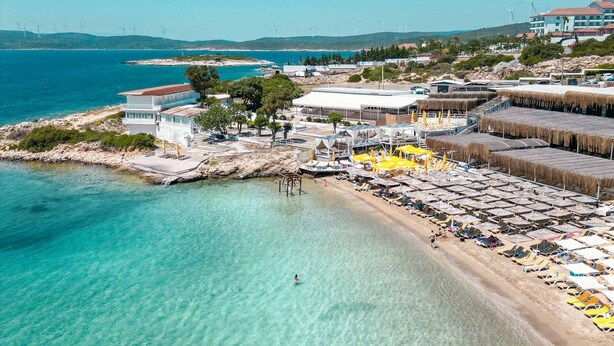 A coastal area with a beach, clear water, and wind turbines in the background. - Lotus City Siğacık Boutique Otel (Seferihisar)