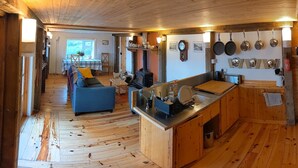 L’intérieur d’un chalet rustique, avec un plancher en bois, un canapé, une table à manger et un coin cuisine où sont suspendues des casseroles.