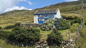 Une maison avec un toit en pente, un mur en pierre et un jardin agrémenté de fleurs et d’arbustes.