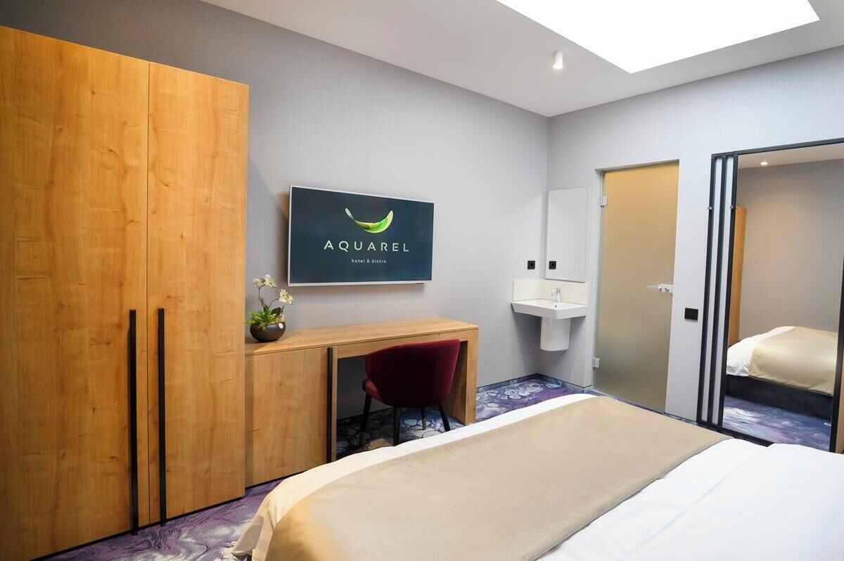 Quarto de hotel moderno com cama, escrivaninha, guarda-roupa, espelho e placa indicativa do Aquarel hotel & bistro.
