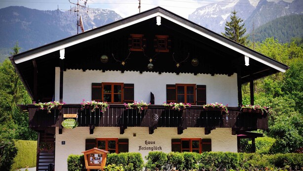 Een klassiek chalet met balkon, omgeven door weelderige begroeiing en bergen op de achtergrond.