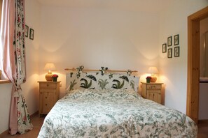 A bedroom with a floral bedspread, two bedside lamps, a wooden headboard, a small wooden nightstand with a vase of flowers, and a window with curtains.