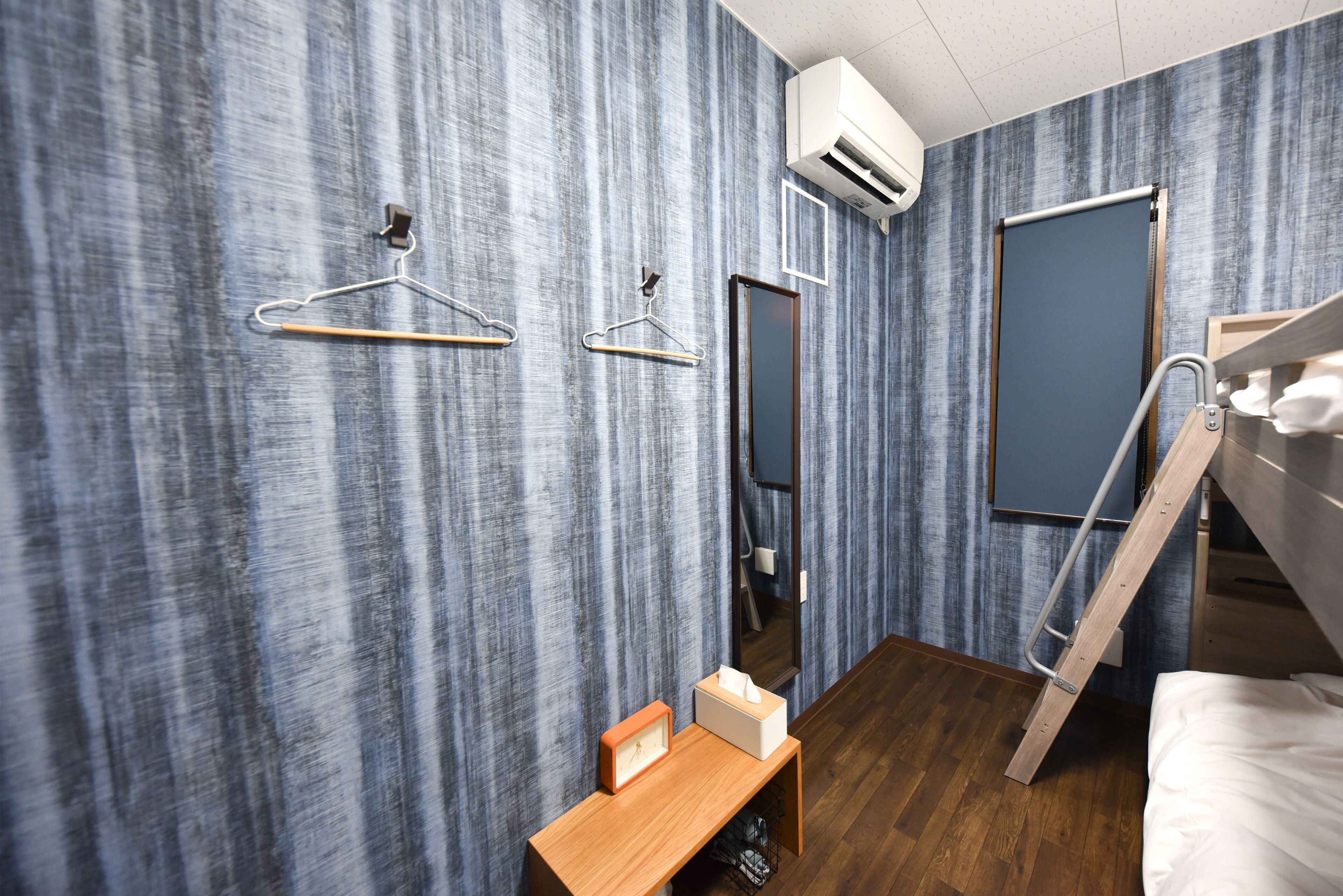 A modern hotel room with a wooden floor, a bunk bed, a wooden desk with a clock and tissues, a mirror, and a wardrobe with a sliding door.