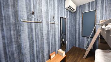 Quarto de hotel moderno com piso de madeira, beliche, escrivaninha de madeira com relógio e lenços, espelho e guarda-roupa com porta deslizante.