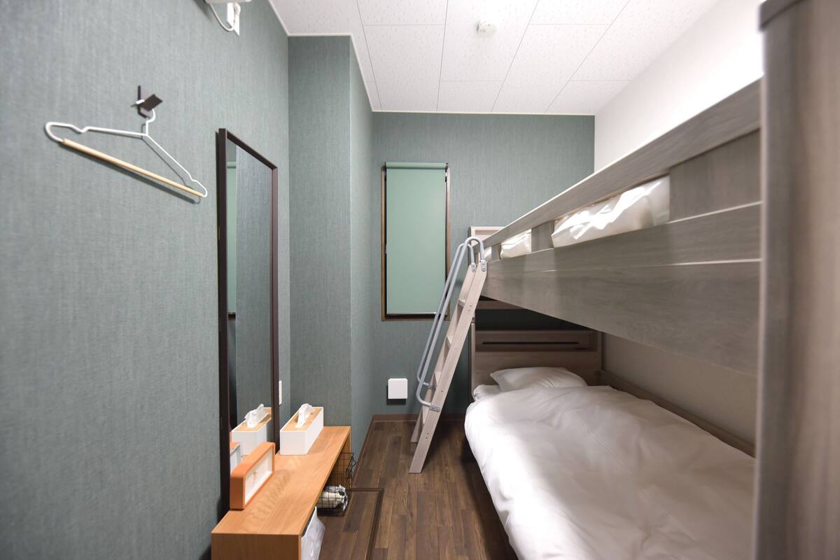 Une chambre d’hôtel compacte comprenant un lit superposé, un bureau en bois avec une boîte de mouchoirs, un miroir, un porte-manteau et une porte.