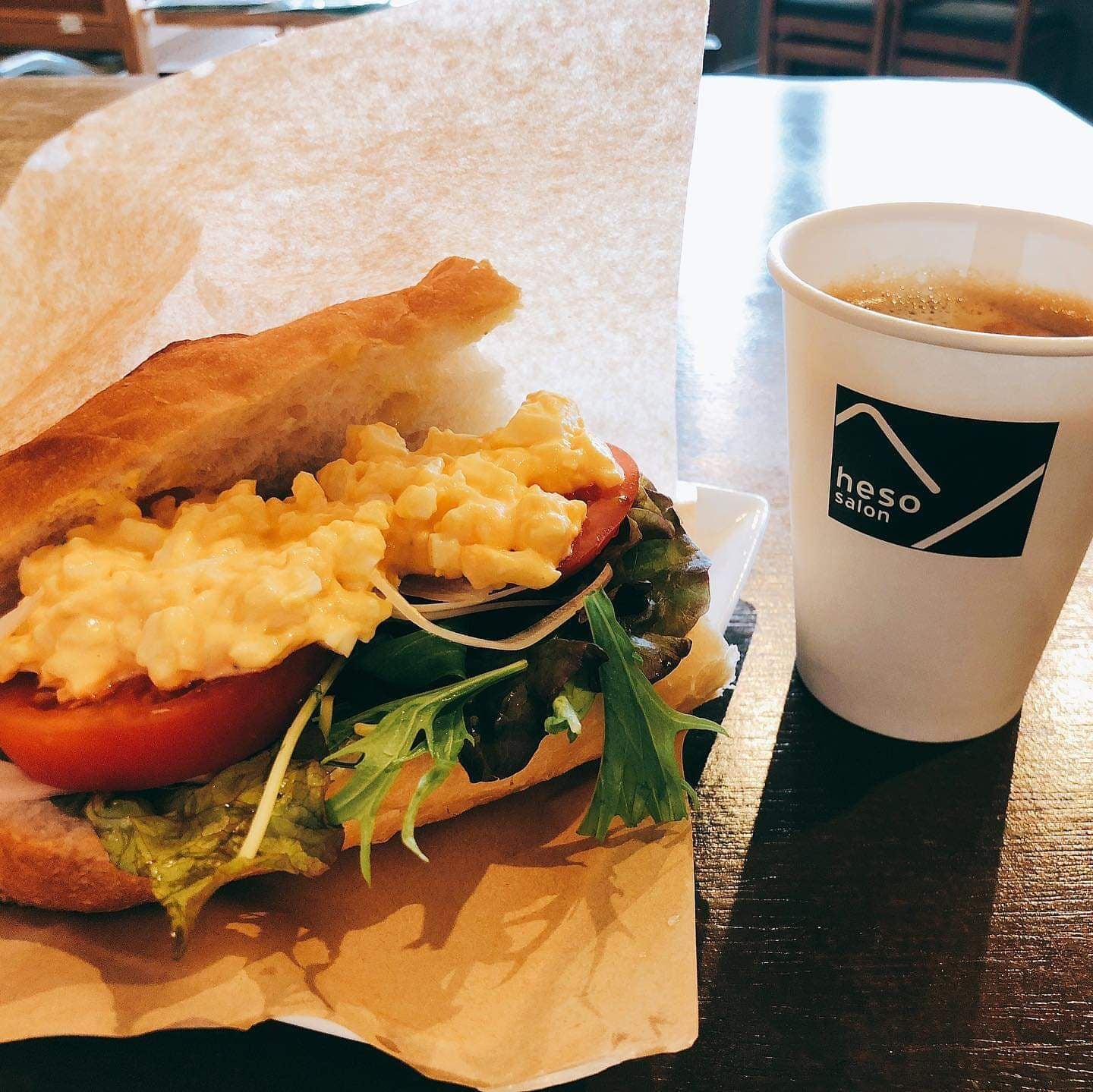 Un sándwich con macarrones y tomate sobre una mesa forrada con papel, junto a una taza con un logotipo.