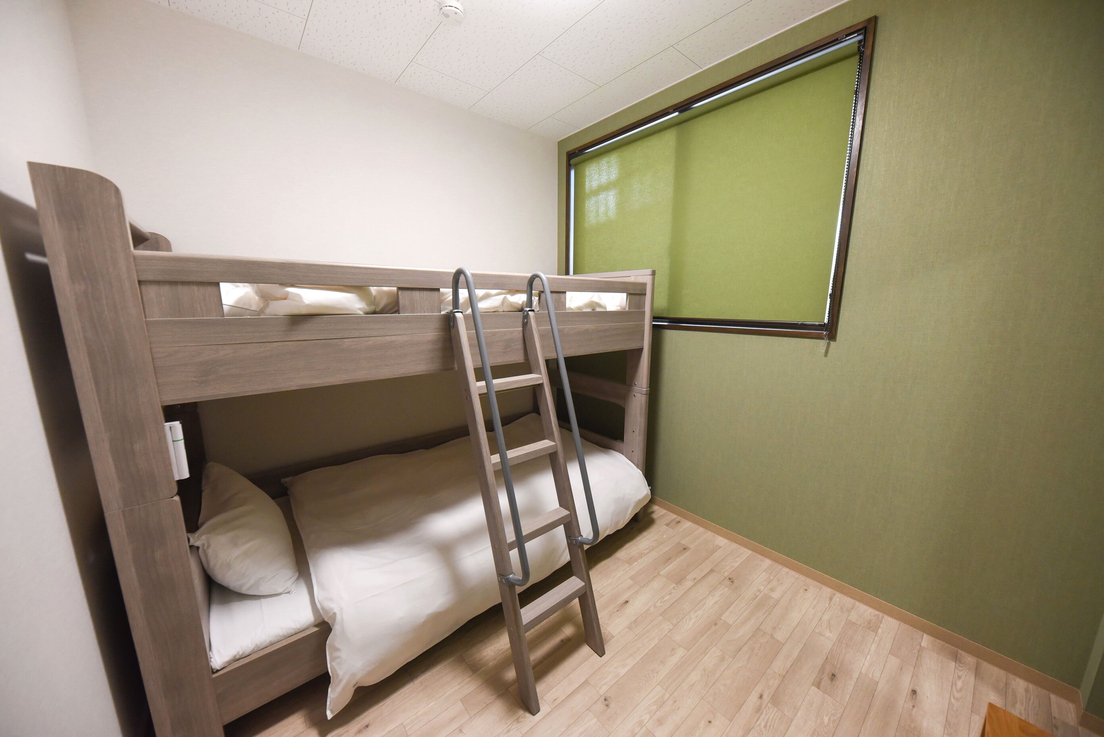 Un lit superposé avec une échelle, une fenêtre avec un store vert et un plancher en bois.