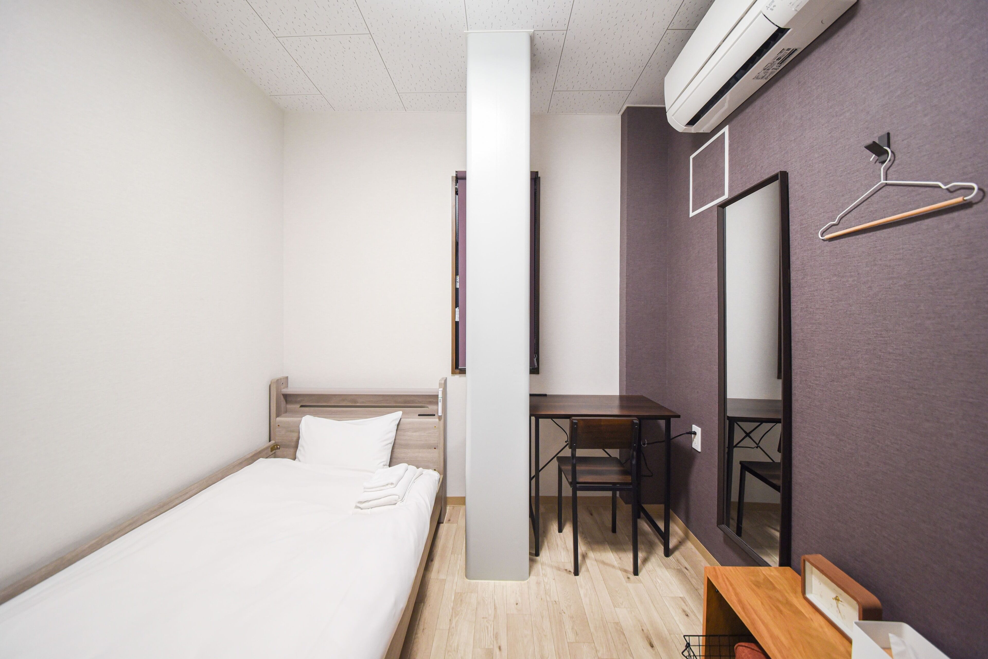 A hotel room with a bed, a desk, a chair, a hanging coat rack, and a mirror on the wall.