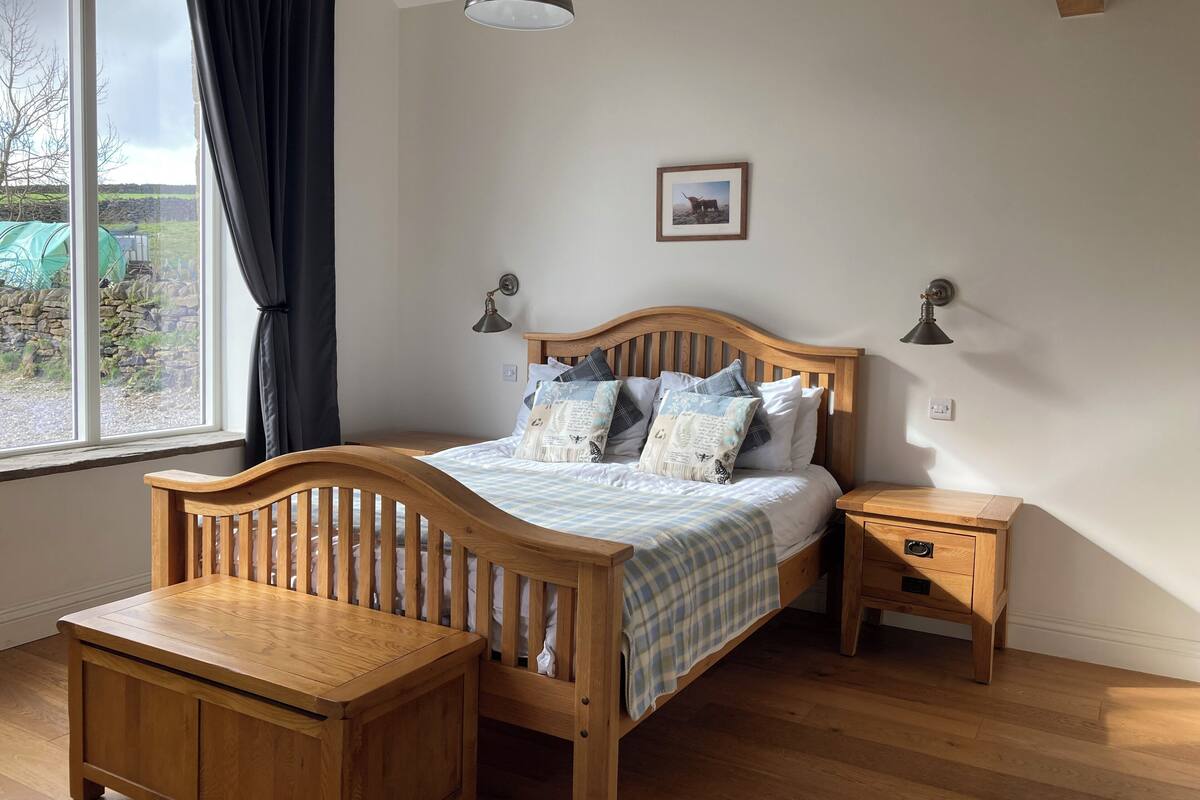 Une chambre à coucher avec un lit en bois, des tables de chevet, un banc en bois, une fenêtre avec des rideaux et une lampe suspendue.