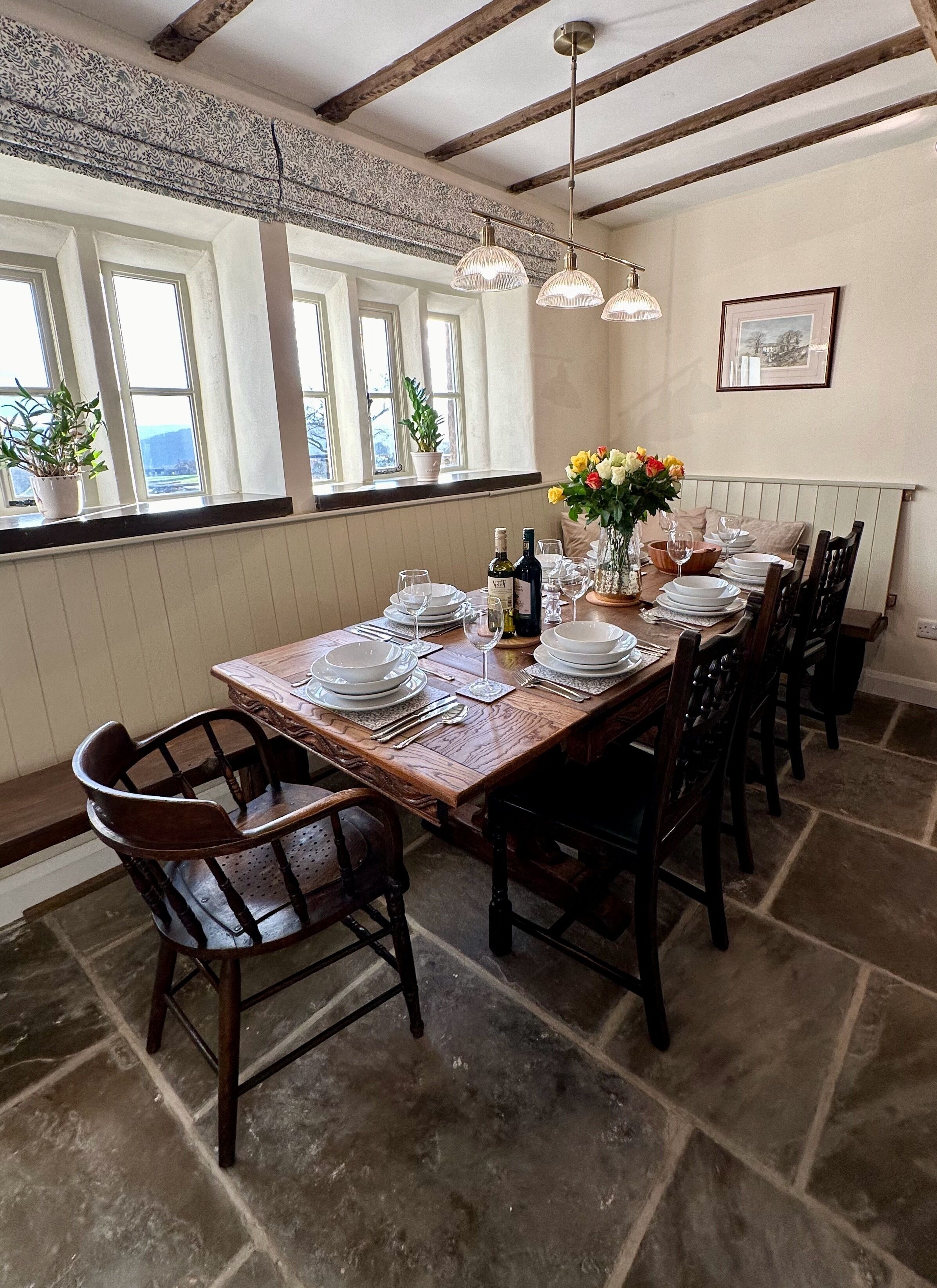 Un comedor con una mesa de madera dispuesta para seis personas, rodeada de sillas. Hay ventanas con plantas en el alféizar, una araña y un cuadro en la pared.