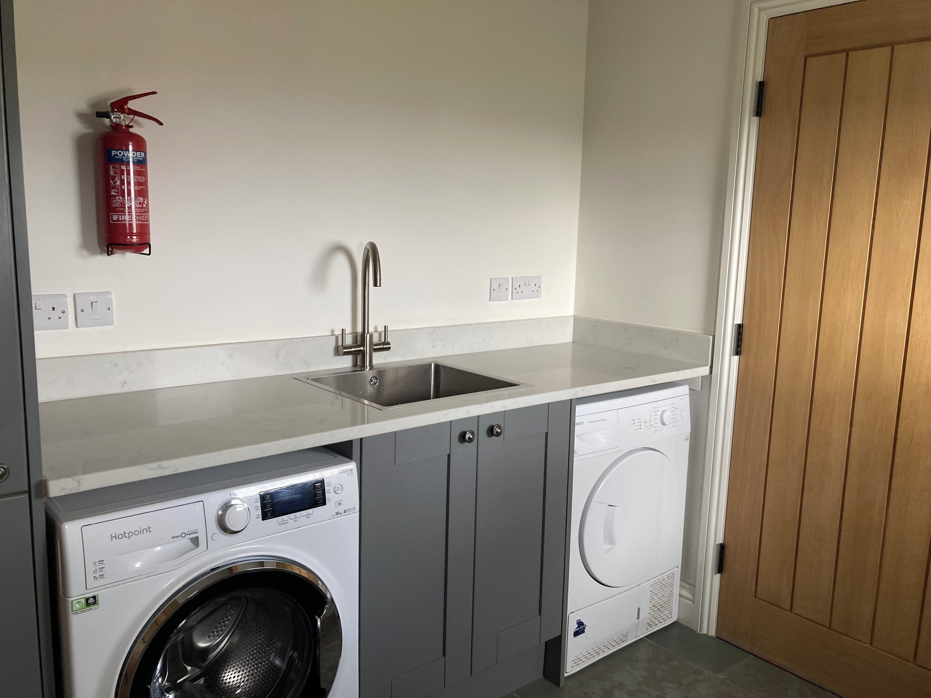 A compact laundry room with a washing machine, a dryer, a stainless steel sink, and a fire extinguisher mounted on the wall.