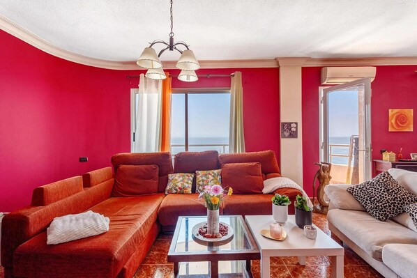 Ein Wohnzimmer mit roter Wand, einer Couch, einem Couchtisch und Blick auf das Meer durch ein großes Fenster.
