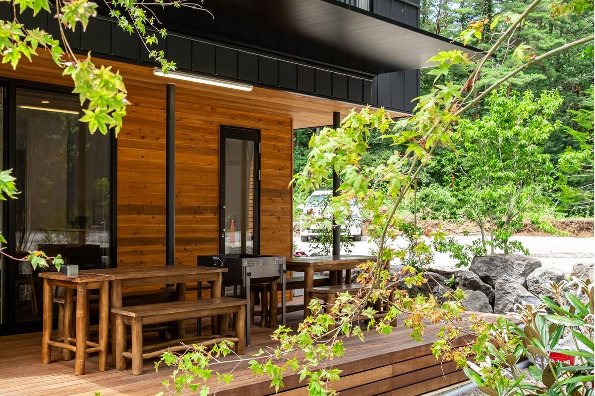 A modern outdoor seating area with wooden tables and benches, surrounded by greenery and rocks.