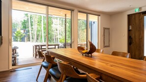 Una mesa de comedor de madera con sillas, ventanas amplias con vista a árboles y una puerta de madera.