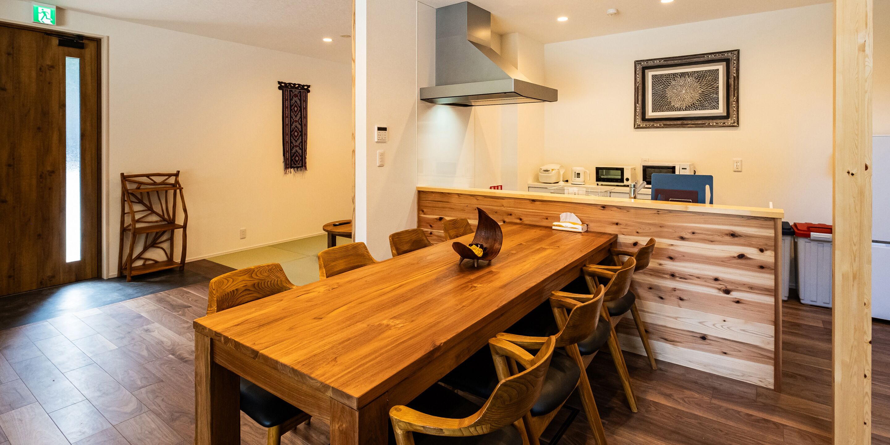 A wooden dining table with chairs, a kitchen area with appliances, and a patterned wall hanging.