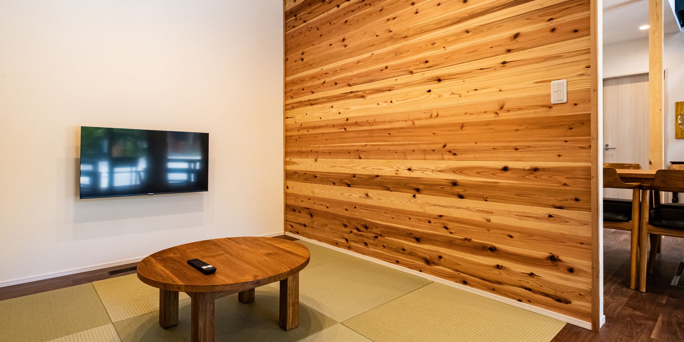 A room with a wooden panel wall, a flat-screen TV mounted on the wall, and a round wooden table with a remote control on it.