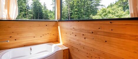 Une cabane en bois avec une grande fenêtre, une baignoire blanche et une vue sur une forêt.