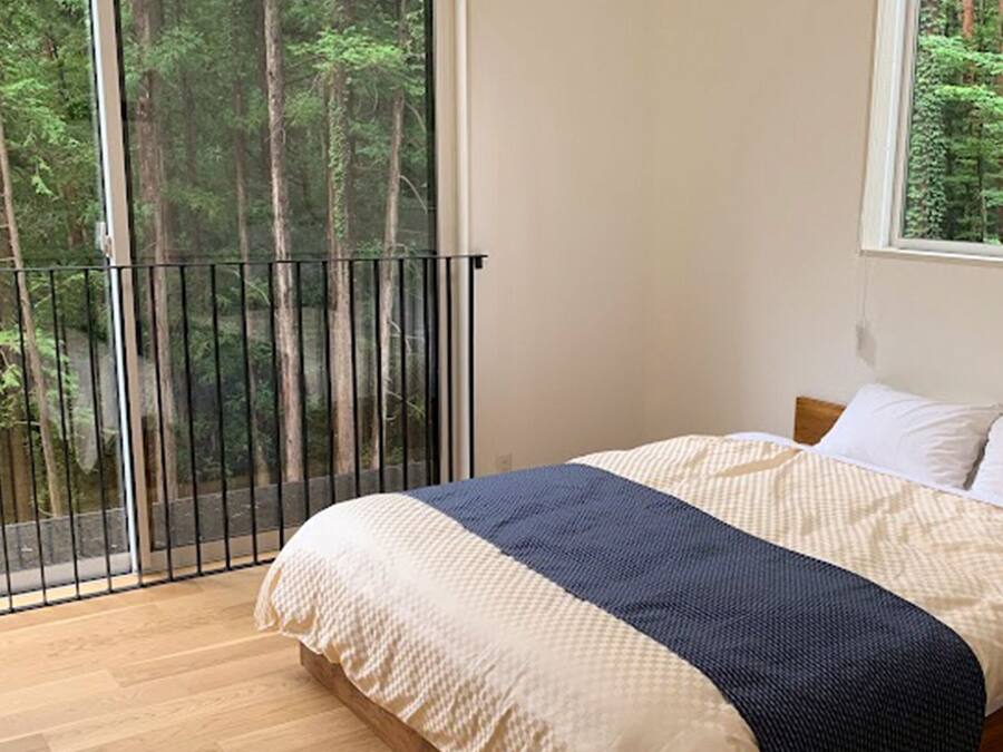 A bedroom with a bed, a window with a forest view, and a balcony with a railing.