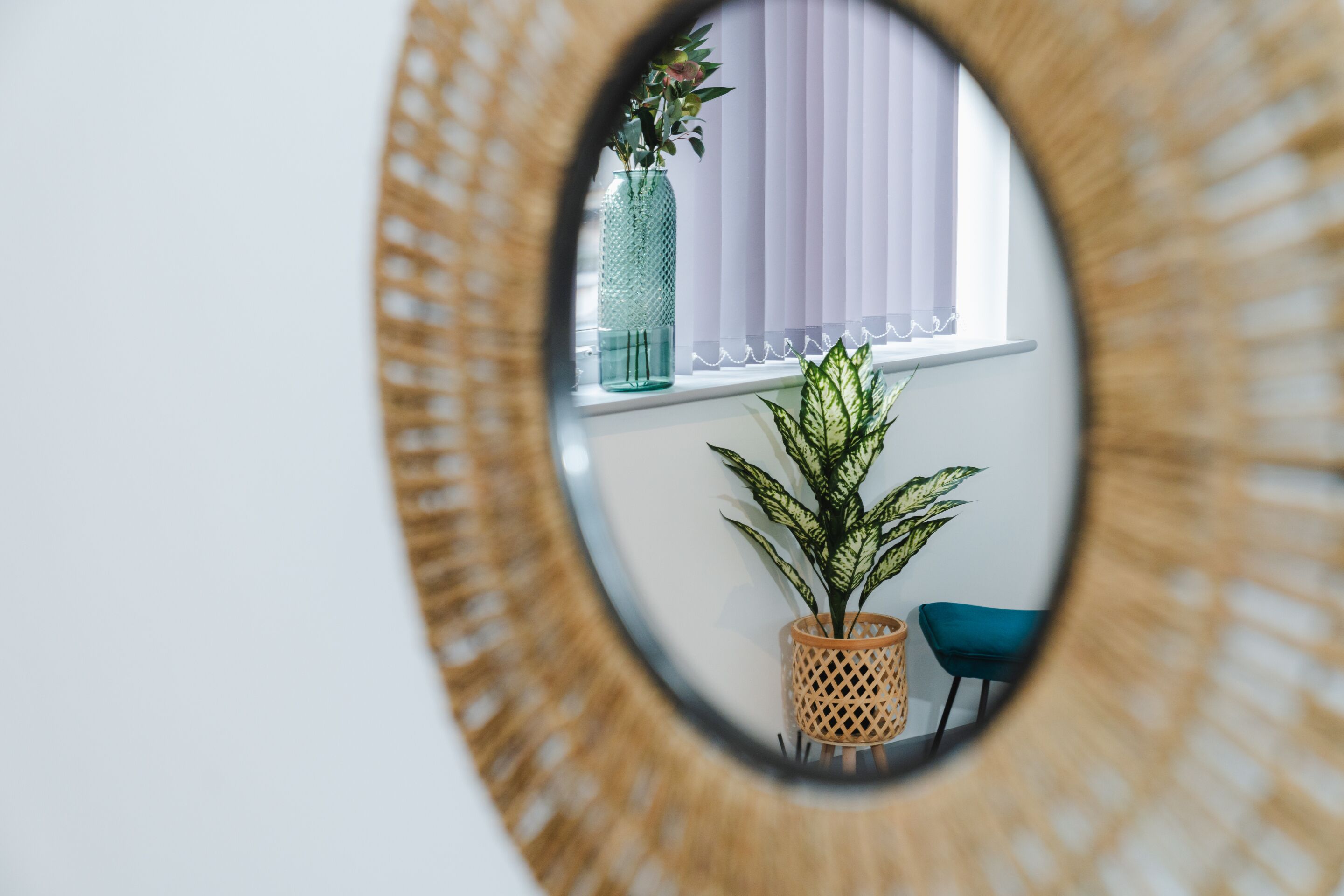 A mirror with a circular frame reflecting a window with blinds, a potted plant, and a chair.
