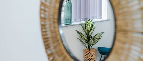 A mirror with a circular frame reflecting a window with blinds, a potted plant, and a chair.