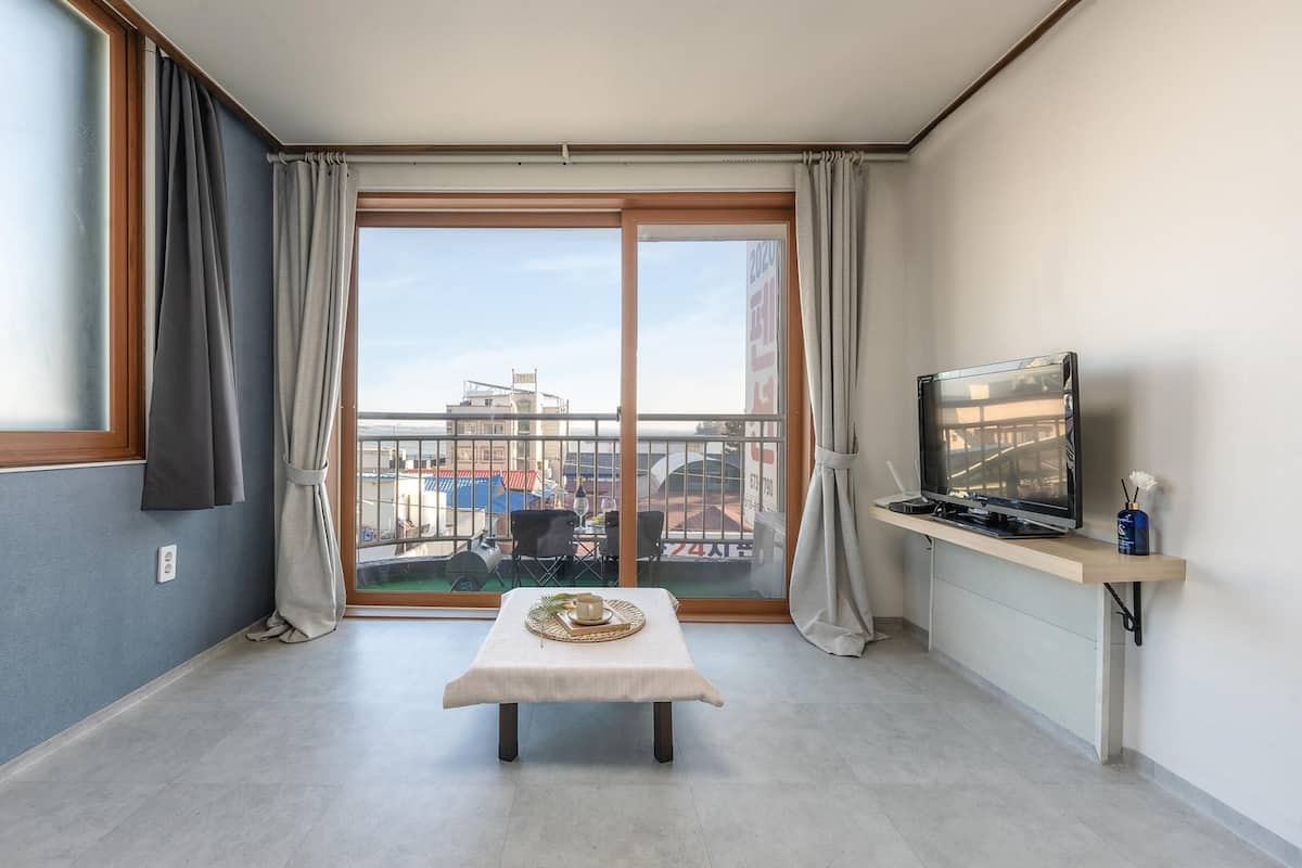 Ein Zimmer mit Schiebeglastür zum Balkon, einem kleinen Tisch mit Schale, einem Fernseher auf einem Schrank und einem Fenster mit Vorhängen.