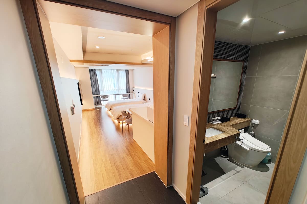 A modern hotel room with a wooden floor, a large bed, a sitting area with a small table and chairs, and a bathroom with a sink and toilet.