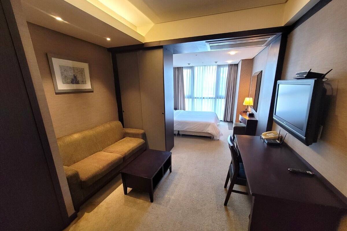 A hotel room with a sofa, a bed, a desk with a lamp, a television, and a picture on the wall.