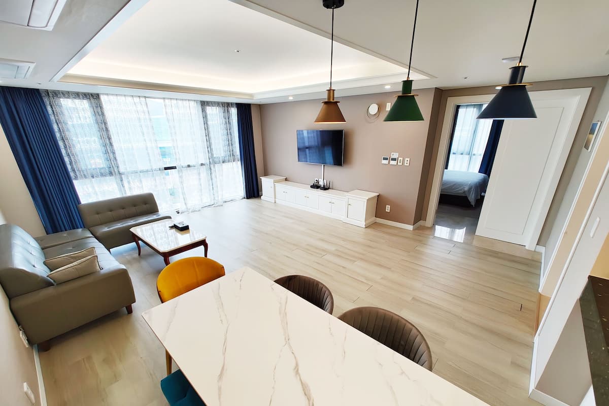 A modern living room with a sofa, coffee table, and a dining area with a marble table. There is a TV mounted on the wall and a window with curtains.
