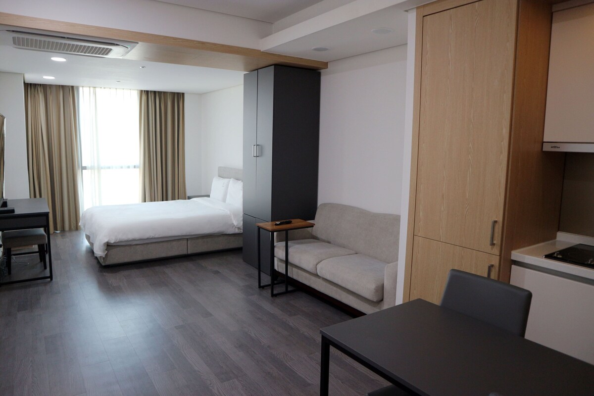 A modern hotel room with a bed, a sofa, a desk, and a kitchenette.