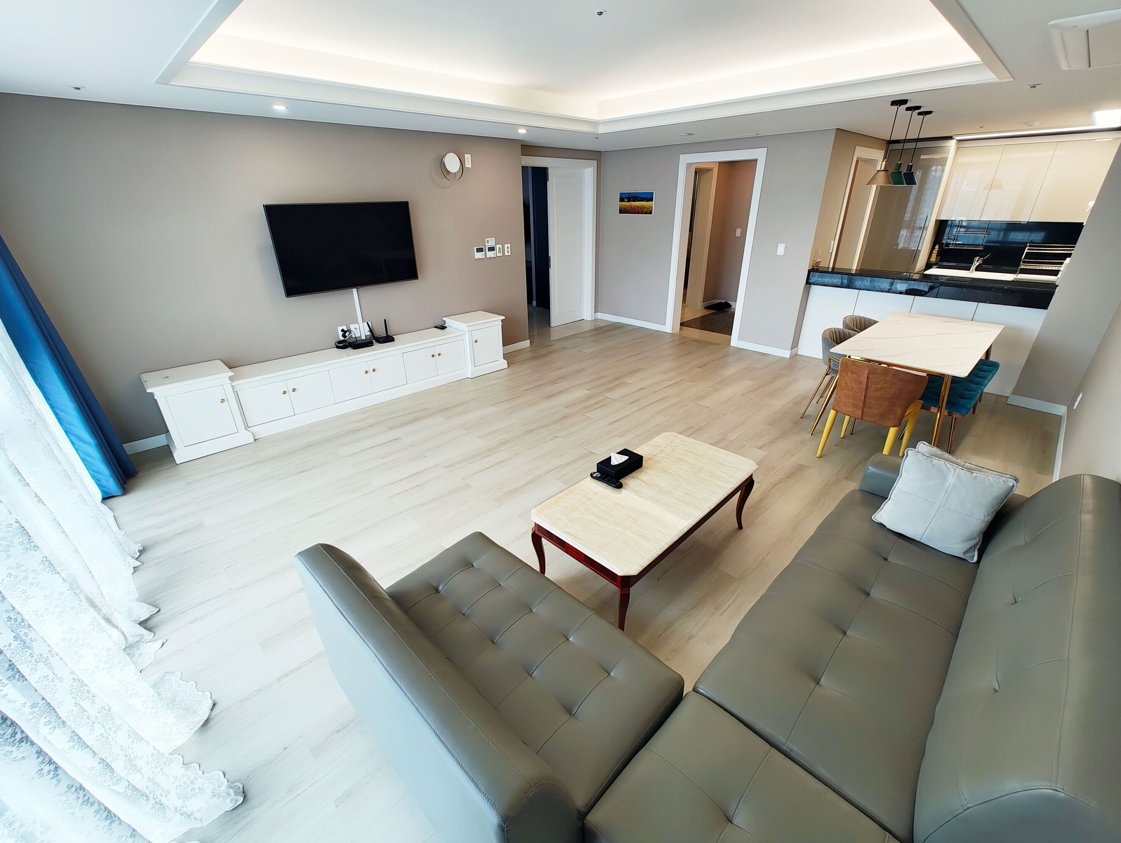 A modern living room with a sofa, a coffee table, and a TV. There is a kitchen area with a dining table and chairs.