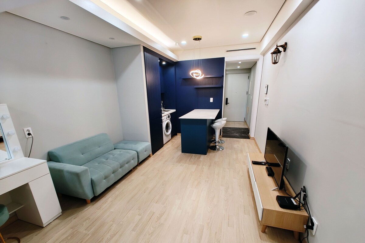 A modern living room with a light blue sofa, a small kitchen area with a washing machine, and a TV on a wooden stand.