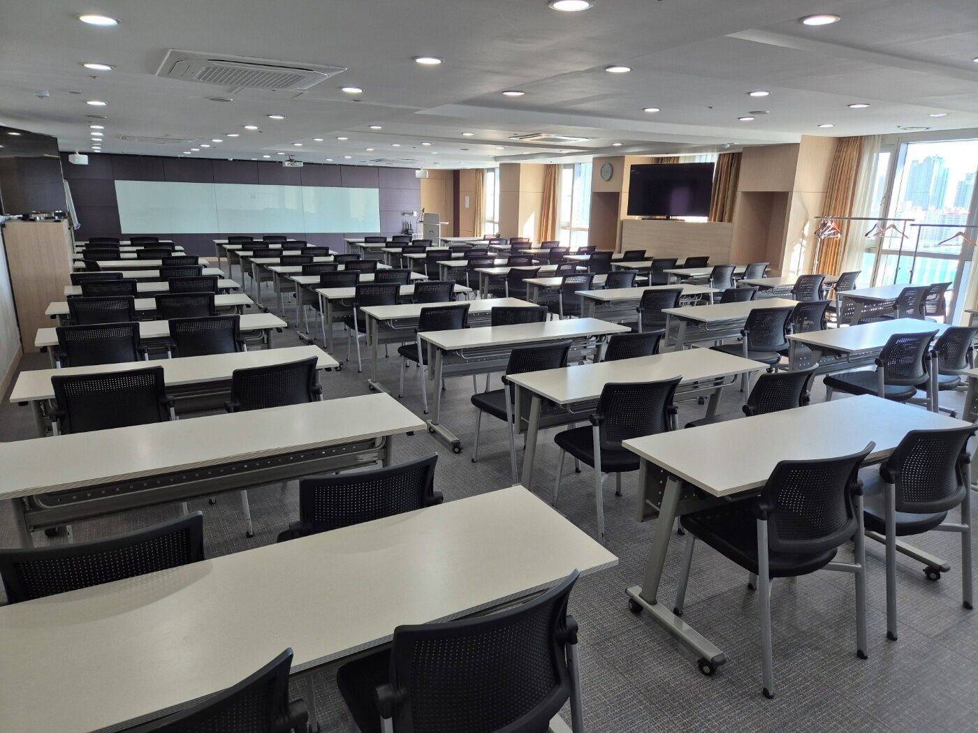 Uma sala de conferências com várias mesas e cadeiras, uma tela grande e uma janela com vista para edifícios.