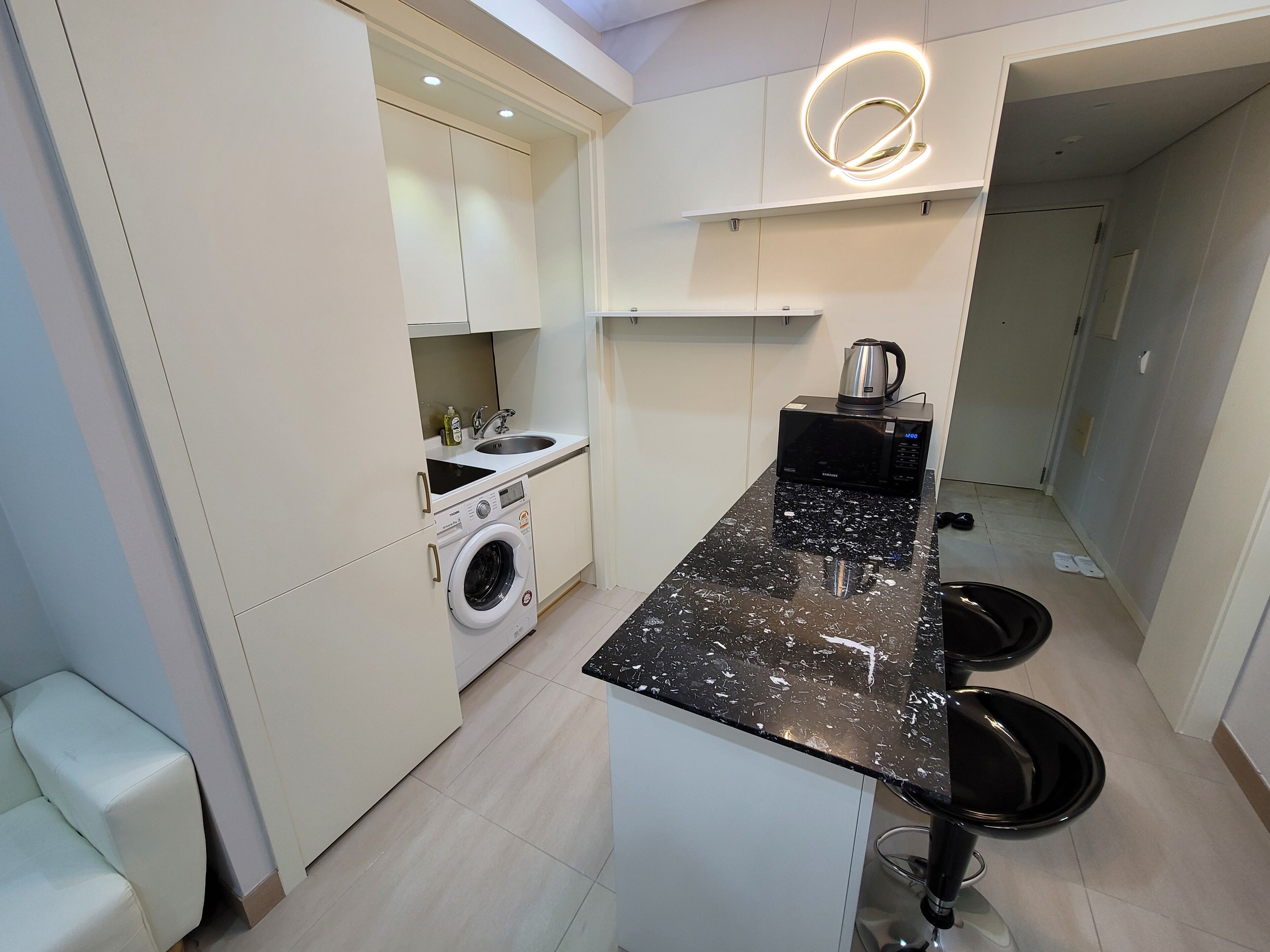 Cocina moderna con lavadora integrada, microondas y pileta. Cuenta con una encimera negra y dos taburetes negros.