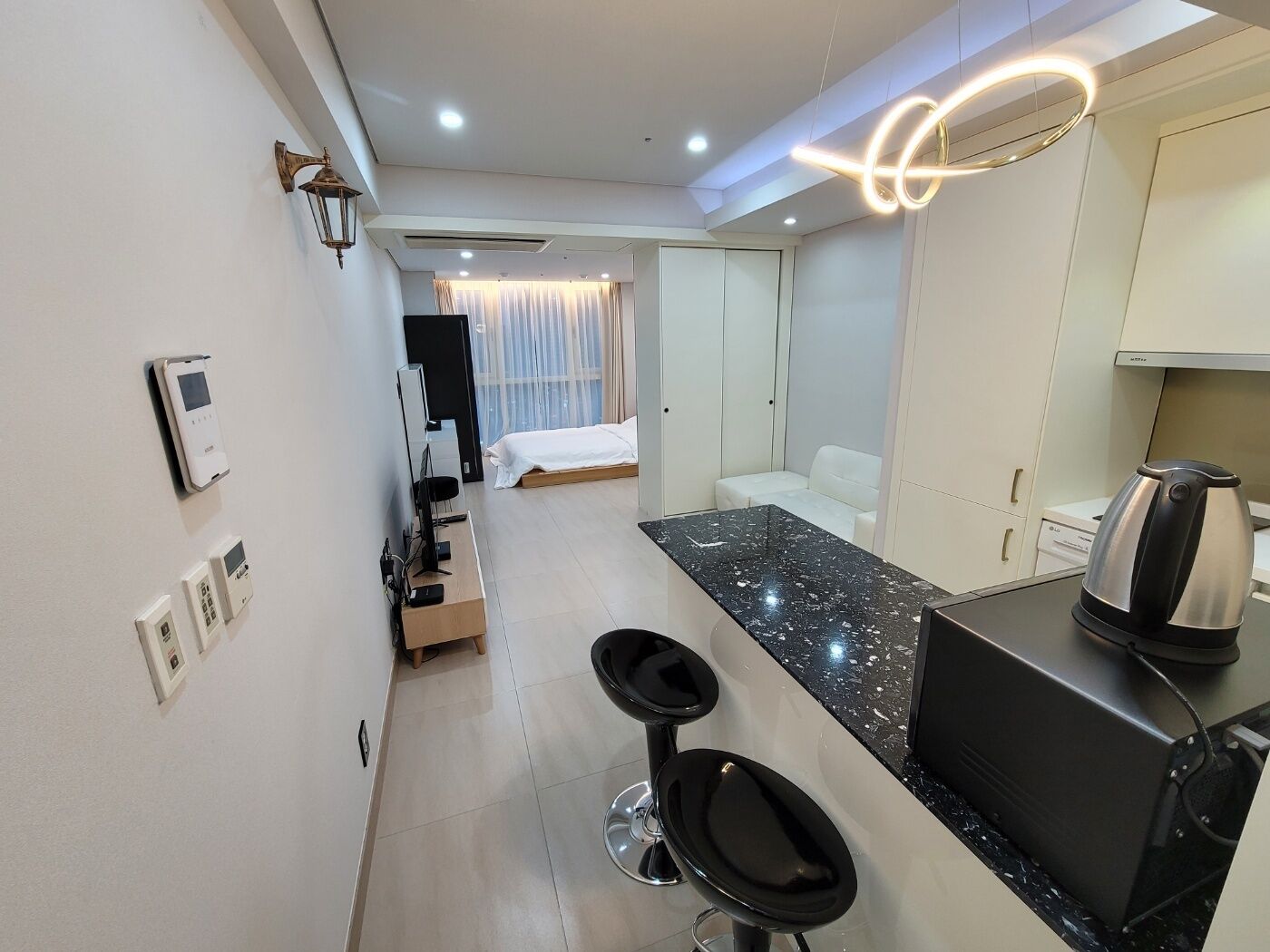 A modern hotel room with a kitchen area, a dining table, and a separate bedroom with a bed and curtains.
