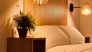 A bedroom with a bed, a bedside table with a plant, a wall-mounted lamp, and a vertical blinds headboard.